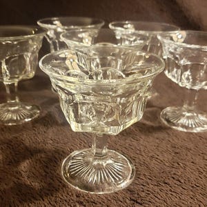 May include: Six clear glass stemmed drinking glasses with a textured, diamond-like pattern. The glasses are arranged in a semi-circle on a brown surface.