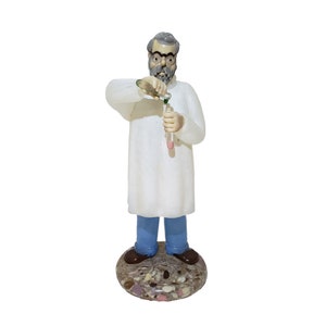 Puede incluir: Una figurita de un científico que lleva una bata de laboratorio blanca y pantalones azules, vertiendo líquido de un vaso de precipitados a un tubo de ensayo. La figurita está de pie sobre una base redonda de mármol marrón y rosa.