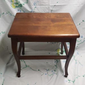 May include: A small, square wooden side table with a rich, warm brown finish. The table features a flat top and curved legs, connected by a lower stretcher for added stability. A classic piece of furniture.