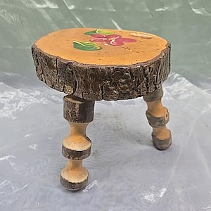 May include: A small, handcrafted wooden stool with a round top and three legs. The top is a natural wood slice, decorated with painted flowers in red, green, and yellow. The legs are light-colored wood with decorative details.