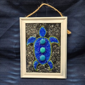 Op de afbeelding: Een ingelijst wandkunstwerk met een blauw zeeschildpadontwerp. De schildpad is gemaakt van getextureerd blauw glas met drie turquoise glazen accenten. Het witte frame heeft een natuurlijke touwlus om op te hangen. De achtergrond is een donker, getextureerd oppervlak.