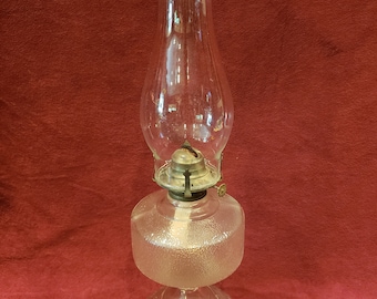 May include: A clear glass oil lamp with a frosted glass base and a decorative etched design. The lamp has a metal wick holder and a clear glass chimney.
