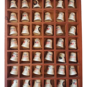 Puede incluir: Una vitrina de madera con 48 pequeños dedalitos de cerámica blanca, cada uno con el retrato y el nombre de un presidente estadounidense diferente. Los dedalitos están dispuestos en forma de cuadrícula.