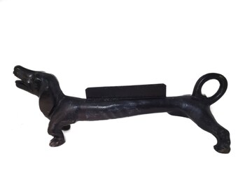 以下が含まれることがあります： 黒い鋳鉄製のダックスフント犬型ドアストッパー。犬は口を開けて立っています。ドアストッパーは背中に長方形のブロックがあり、尻尾の端にループが付いています。