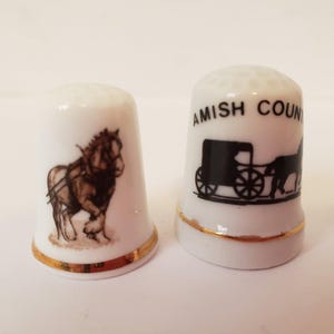 Può includere: Due ditale in ceramica bianca con bordatura dorata. Un ditale presenta un'illustrazione in bianco e nero di un cavallo che tira una carrozza con il testo "Amish Country". L'altro ditale presenta un'illustrazione in bianco e nero di un cavallo.