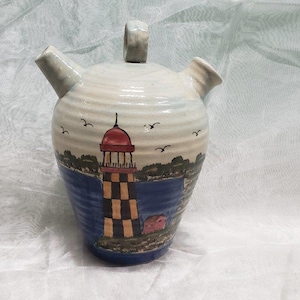 May include: A ceramic pitcher with a lighthouse design. The pitcher is white with blue and green accents. It has a red roof and a black and white striped tower. The pitcher has three spouts.