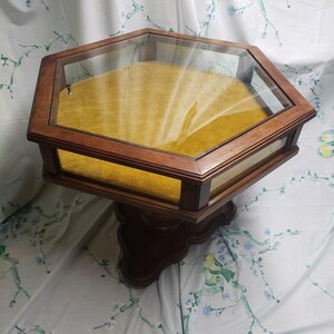 May include: A vintage display case with a hexagonal glass top and a wooden frame. The interior is lined with yellow fabric. The case is supported by a decorative wooden base. The overall color scheme is brown and yellow.
