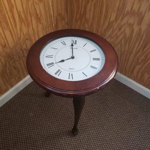 May include: A round, dark brown wooden side table with a clock face. The clock has Roman numerals and black hands. The table has three curved legs and is on a brown carpet.