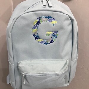 May include: A light blue backpack with a front pocket and a zip closure. The backpack has a dinosaur-shaped letter "G" embroidered on the front.