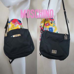May include: Black nylon shoulder bag with gold hardware and the word "MOSCHINO" in gold letters. The bag has a long adjustable strap and a zipper closure.