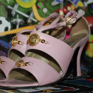 May include: A pair of light pink high-heeled sandals. The sandals feature gold-colored embellishments and a strap that buckles around the ankle. The heels are approximately 4 inches tall.