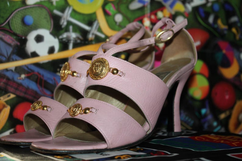 May include: A pair of light pink high-heeled sandals. The sandals feature gold-colored embellishments and a strap that buckles around the ankle. The heels are approximately 4 inches tall.