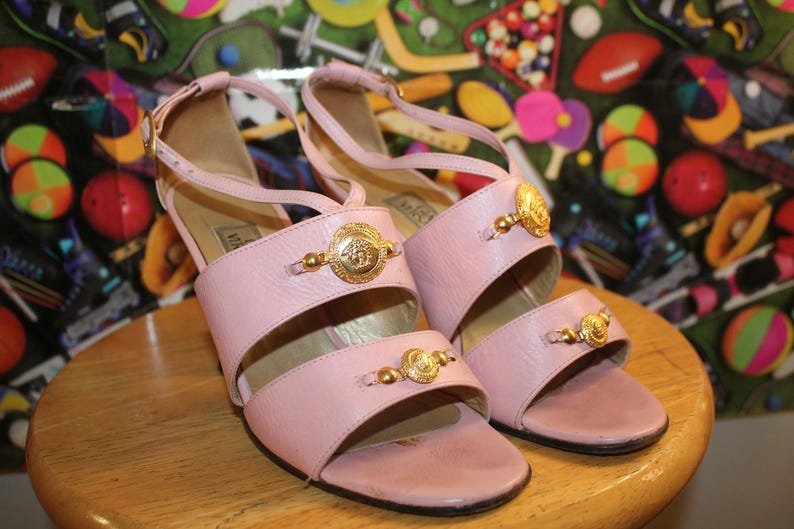 May include: A pair of light pink leather sandals with gold-tone Medusa head accents. The sandals have a strappy design with a small heel and are displayed on a wooden surface.
