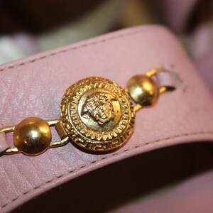 May include: Close-up of a light pink leather sandal strap. The strap features a gold-colored medallion with a detailed design and two smaller gold accents. The sandal's design is classic and elegant.