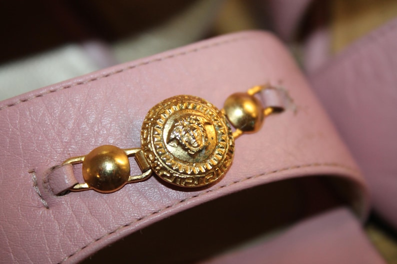 May include: Close-up of a light pink leather sandal strap. The strap features a gold-colored medallion with a detailed design and two smaller gold accents. The sandal's design is classic and elegant.