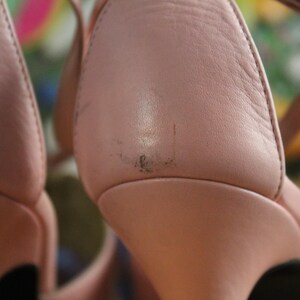 May include: Close-up of a pair of pink leather high-heeled sandals. The sandals have a strap around the ankle and a gold-colored buckle. The leather shows some wear and tear.