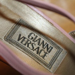 May include: Close-up of a pink leather shoe interior with a gold-colored insole. The label reads "GIANNI VERSACE" in black text within a black and white border. The shoe's strap is visible.
