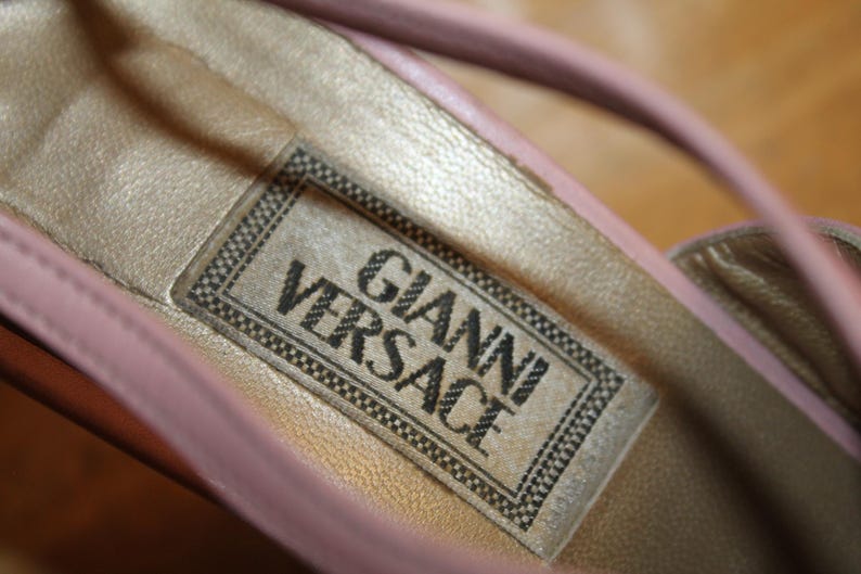 May include: Close-up of a pink leather shoe interior with a gold-colored insole. The label reads "GIANNI VERSACE" in black text within a black and white border. The shoe's strap is visible.