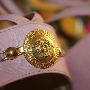 May include: Close-up of a pair of pink leather sandals. The sandals feature a gold-colored Medusa head emblem and gold hardware. The sandals have a strap design.