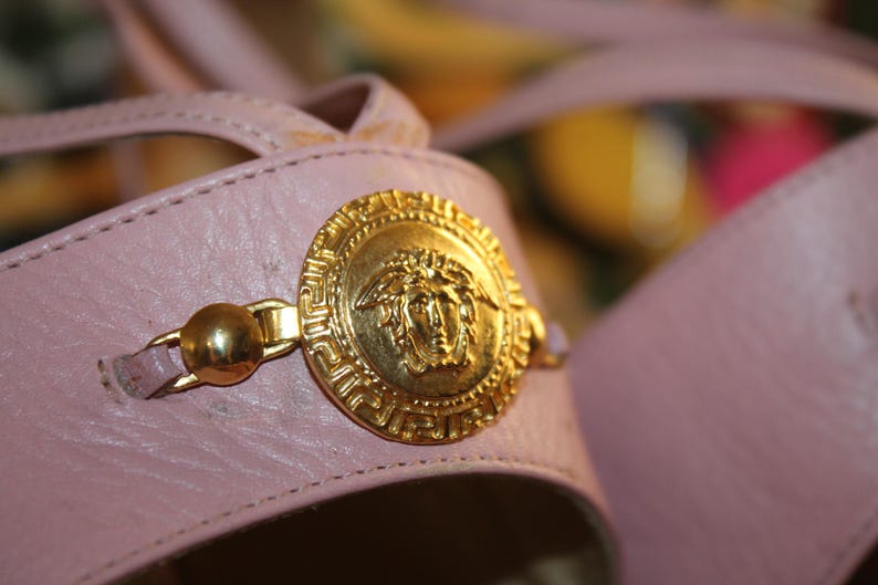 May include: Close-up of a pair of pink leather sandals. The sandals feature a gold-colored Medusa head emblem and gold hardware. The sandals have a strap design.