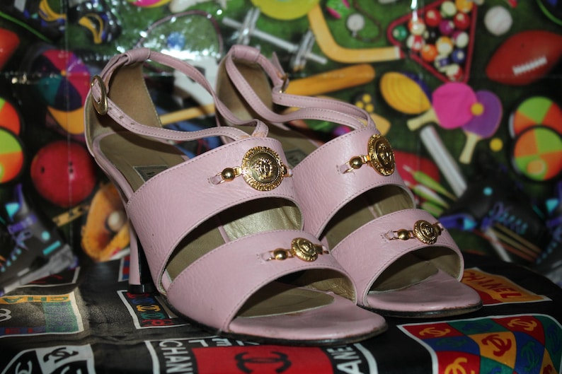 May include: A pair of light pink high-heeled sandals. The sandals feature gold-colored embellishments and a strap around the ankle. The heels are approximately 3 inches tall. The sandals are displayed on a colorful patterned surface.