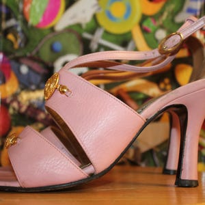 May include: A light pink sandal with a high heel. The sandal features a strap across the toes and a strap around the ankle, with a gold-colored buckle. The heel is approximately 4 inches (10 cm) high, and the sandal has gold-colored embellishments.