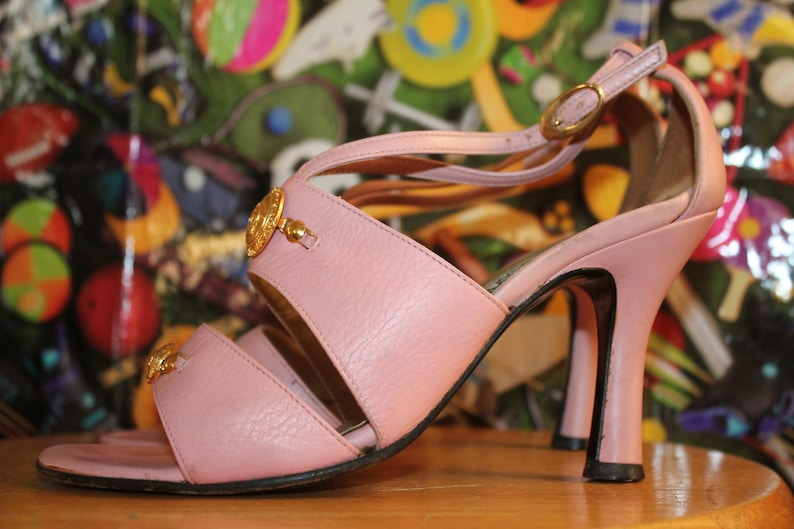May include: A light pink sandal with a high heel. The sandal features a strap across the toes and a strap around the ankle, with a gold-colored buckle. The heel is approximately 4 inches (10 cm) high, and the sandal has gold-colored embellishments.