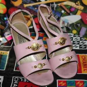 May include: A pair of light pink leather sandals with gold-tone Medusa head accents. The sandals have a strappy design and a high heel. The background features a colorful pattern and a black and white Chanel logo.