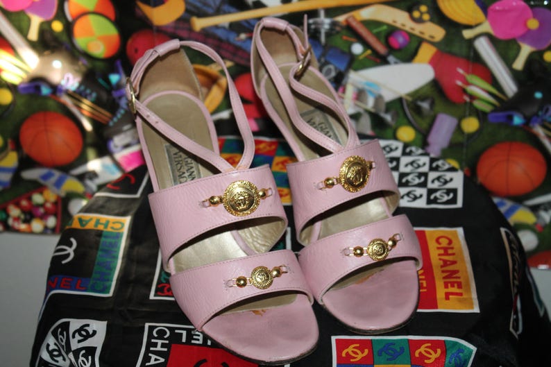 May include: A pair of light pink leather sandals with gold-tone Medusa head accents. The sandals have a strappy design and a high heel. The background features a colorful pattern and a black and white Chanel logo.