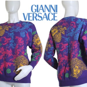 May include: Purple sweater with a crew neck and long sleeves. The sweater features a colorful pattern of floral and Medusa head designs in yellow, pink, blue, and green. The text "GIANNI VERSACE" is displayed in blue.