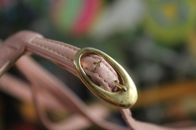 May include: Close-up of a pale pink leather strap with a gold-colored oval buckle. The strap is stitched and appears to be part of a larger accessory, possibly a handbag or shoe.
