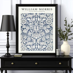 Puede incluir: Una impresi&oacute;n enmarcada en negro con un patr&oacute;n floral blanco sobre un fondo azul. La impresi&oacute;n dice "WILLIAM MORRIS - COTTON PRINTS EXHIBITION". La impresi&oacute;n se muestra en una consola negra con una l&aacute;mpara negra y un jarr&oacute;n con ramas verdes.
