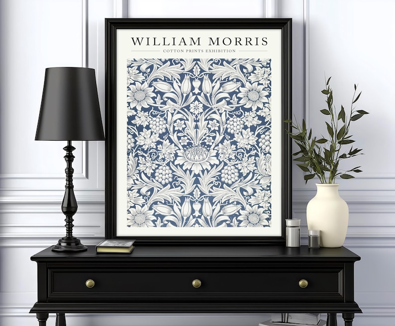 Puede incluir: Una impresi&oacute;n enmarcada en negro con un patr&oacute;n floral blanco sobre un fondo azul. La impresi&oacute;n dice "WILLIAM MORRIS - COTTON PRINTS EXHIBITION". La impresi&oacute;n se muestra en una consola negra con una l&aacute;mpara negra y un jarr&oacute;n con ramas verdes.