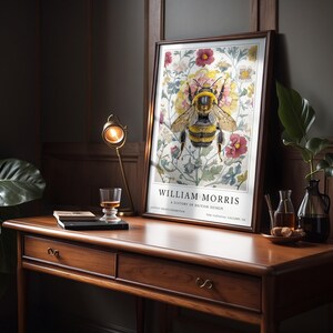 May include: A framed print of a bee surrounded by flowers. The print is titled "William Morris: A History of British Design." The print is on a wooden desk with a glass of liquid and a book.