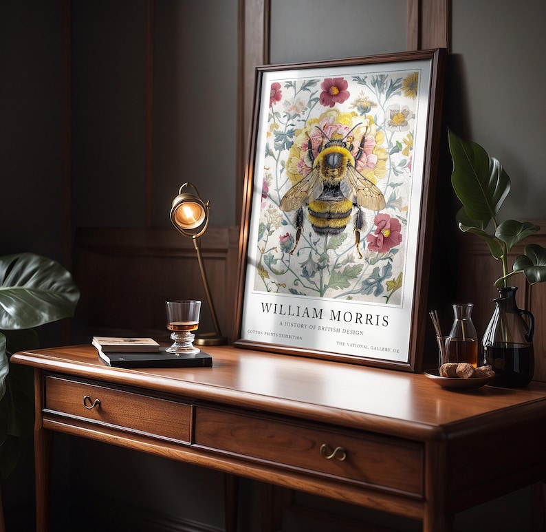 May include: A framed print of a bee surrounded by flowers. The print is titled "William Morris: A History of British Design." The print is on a wooden desk with a glass of liquid and a book.