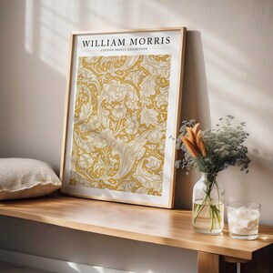 May include: A framed print of a yellow and white floral pattern with the text "WILLIAM MORRIS COTTON PRINTS EXHIBITION" at the top. The print is hanging on a wall above a wooden shelf.