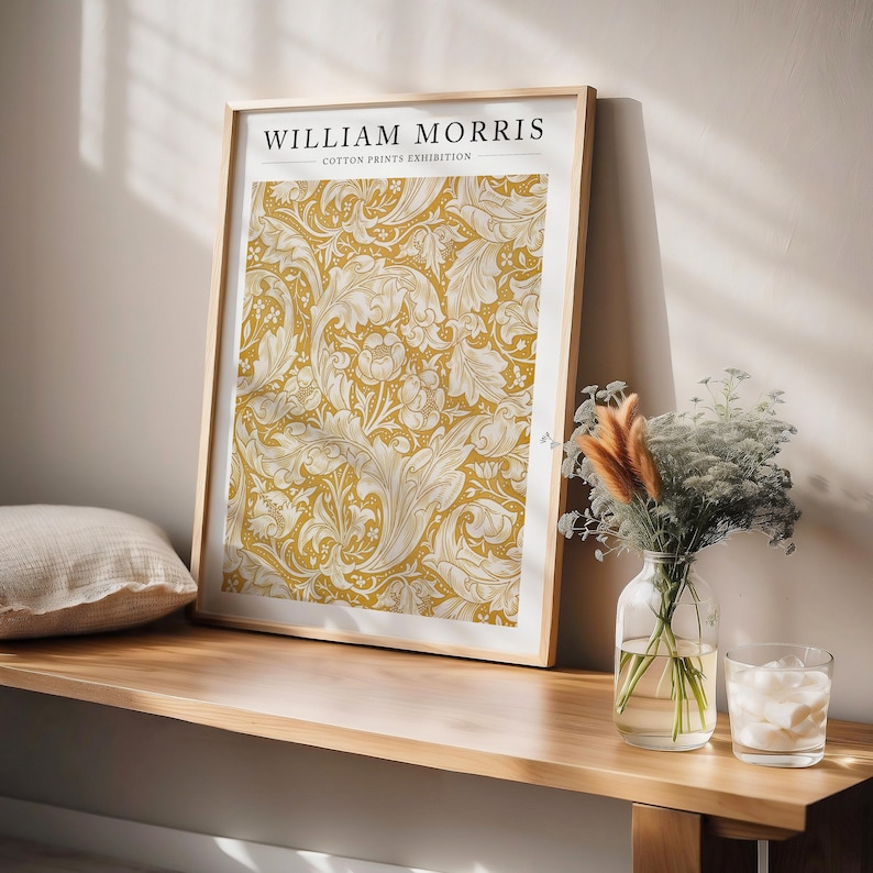 May include: A framed print of a yellow and white floral pattern with the text "WILLIAM MORRIS COTTON PRINTS EXHIBITION" at the top. The print is hanging on a wall above a wooden shelf.