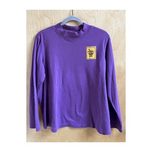May include: A purple long-sleeved shirt with a mock neck and a small yellow patch on the left chest. The patch features a brown illustration of a cup with a spoon.
