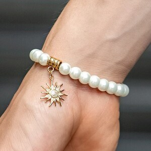 May include: A white pearl bracelet with a gold sun charm. The bracelet is strung on an elastic band and features a gold clasp. The sun charm is detailed with small crystals. The bracelet is worn on a wrist.