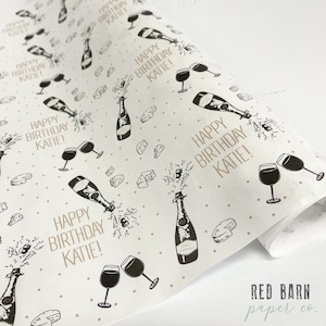 May include: White wrapping paper with a repeating pattern of champagne bottles popping open, wine glasses, and small pieces of cheese. The text "Happy Birthday Katie!" is repeated throughout the pattern.