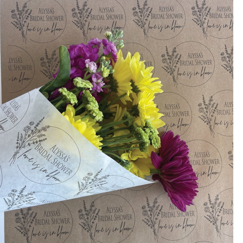 May include: A bouquet of yellow, pink, and purple flowers wrapped in white tissue paper with a brown paper background. The brown paper has repeating circles with the text "Alyssa's Bridal Shower Love is in Bloom".