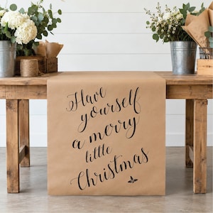 May include: A brown kraft paper table runner featuring the black printed text Have yourself a merry little Christmas in a cursive script font, with a small holly leaf icon positioned below the final word of the phrase.