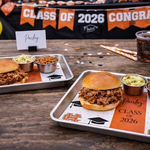 May include: Two metal trays with pulled pork sandwiches, coleslaw, and baked beans. The trays have a graduation theme with the text "Paisley CLASS OF 2026". A drink and decorations are in the background.