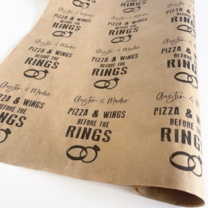 May include: Brown paper gift wrap with the text "Austin & Madie Pizza & Wings Before the Rings" repeated in a pattern. The text is in a black font with a distressed look. The design also includes a ring graphic.