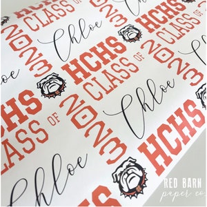May include: White wrapping paper with a repeating pattern of "HCHS CLASS OF 2023" in orange, along with the name "Chloe" in a cursive font. The design includes a bulldog mascot logo.