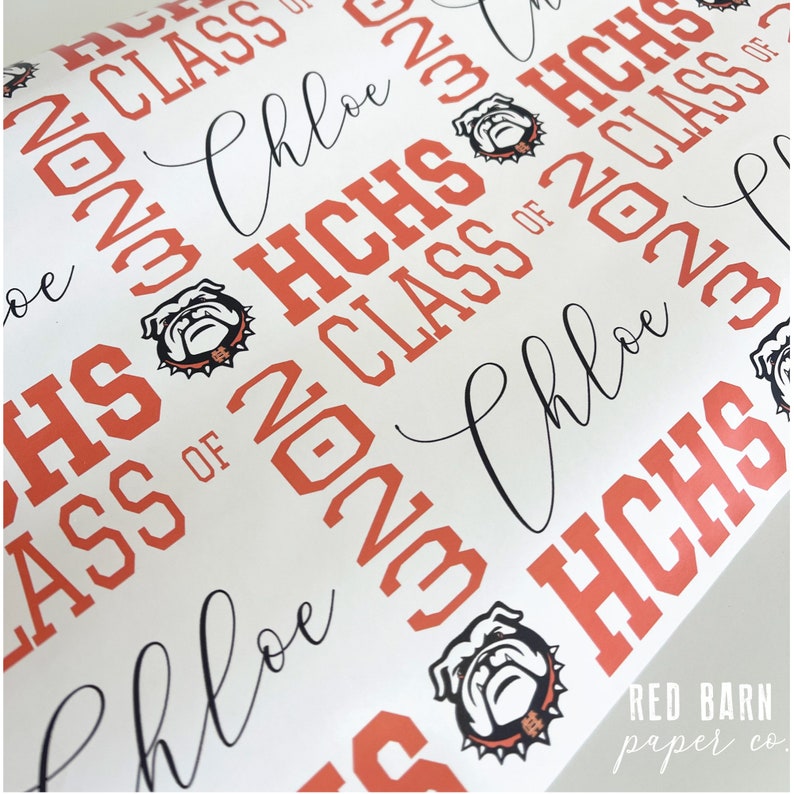 May include: White wrapping paper with a repeating pattern of "HCHS CLASS OF 2023" in orange, along with the name "Chloe" in a cursive font. The design includes a bulldog mascot logo.