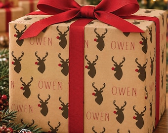 May include: A square gift box wrapped in light brown kraft paper featuring a repeating pattern of dark brown reindeer silhouettes with red noses and the name OWEN printed in red. The box is tied with a wide, solid red grosgrain ribbon bow.