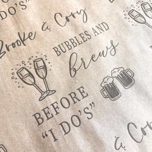 May include: A brown paper bag with the text "Brooke & Cory" and "Bubbles and Brews Before "I Do's"" printed on it. The bag also features illustrations of champagne flutes and beer mugs.