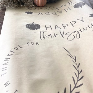 May include: A beige table runner with the text "Happy Thanksgiving" and "I am thankful for" printed in black. The runner is decorated with a black leaf design and a small pumpkin silhouette.
