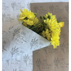 May include: A bouquet of yellow flowers wrapped in white tissue paper with a repeating pattern of black and white floral designs and the text "Baby O'Brien in Bloom".